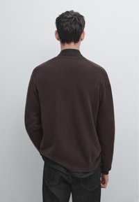 Brauner Strickpullover mit Rundhalsausschnitt und Raglanärmeln, der eine weiche Textur aufweist, kombiniert mit dunklen Jeans, von hinten betrachtet.