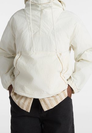 Veste pullover blanche légère avec une finition texturée, capuche réglable, poches latérales et coutures contrastées. Portée sur une chemise à motifs.