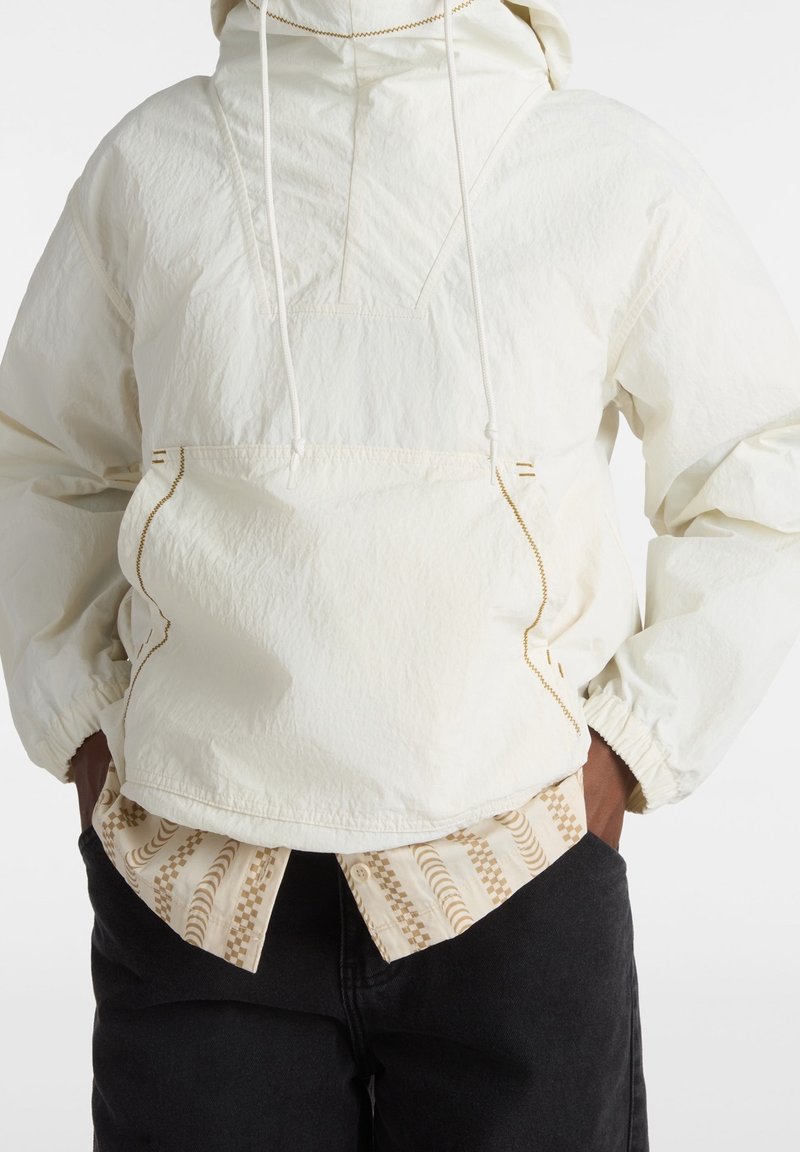 Lättviktig vit pulloverjacka med en strukturerad yta, justerbar huva, sidofickor och kontrasterande sömmar. Burens över en mönstrad skjorta.