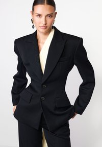 Blazer negru croit, cu umeri structurați, două butoane �și buzunare frontale. Purtați peste un top plisat de un galben deschis. Textură de material neted.