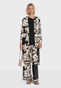 Tenue à motif floral noir et crème comprenant un long cardigan ouvert et un pantalon à jambes larges, complétée par un haut noir uni et un petit sac.