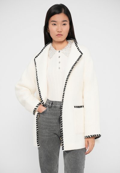 Veste en polaire blanche avec des finitions noires, design sans col, et poche avant, associée à un top en tricot côtelé et un jean taille haute gris.
