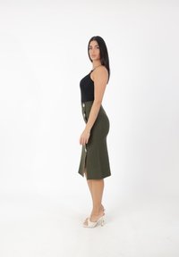 Top nero senza maniche abbinato a una gonna midi fitted verde oliva con uno spacco frontale e bottoni bianchi al centro. Indossato con tacchi bianchi.
