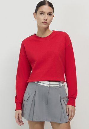 Un sweat-shirt rouge à manches longues associé à une jupe plissée grise. La jupe présente une ceinture blanche et des poches, mettant en valeur une finition texturée.