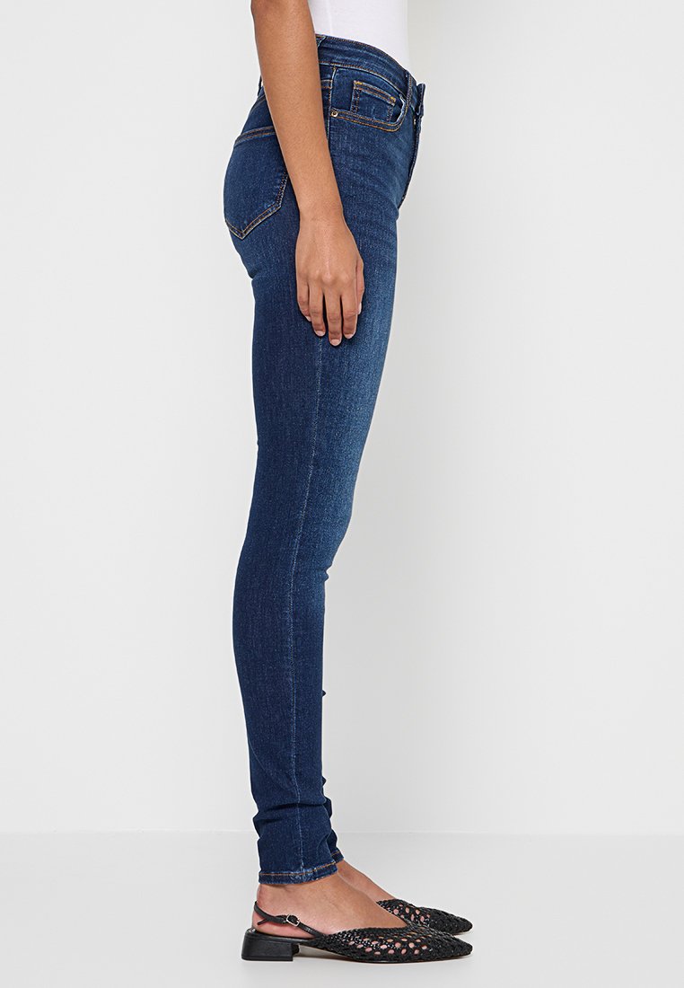 Dunkelblaue Skinny-Jeans mit hoher Taille, aus Denim-Stoff gefertigt. Verfügt über Gesäßtaschen und eine glatte Textur. Kombiniert mit schwarzen Schuhen.