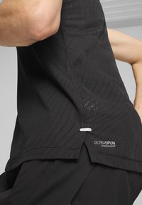 Juodas sportinis topas, pagamintas iš perforuoto audinio, su subtilia tekstūra, išlenkta apačia ir atspindinčiu žyma "ULTRASPUN" šalia siūlės.