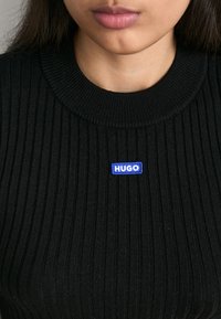 Pull noir côtelé avec un col rond et un patch logo rectangulaire bleu portant l'inscription "HUGO" centré sur la poitrine.
