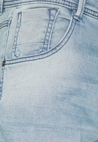 Lichtblauwe denim jeans hebben een gestructureerde pasvorm, een voorkant zak en zichtbare stikselaccenten. De stof lijkt iets gestructureerd te zijn.