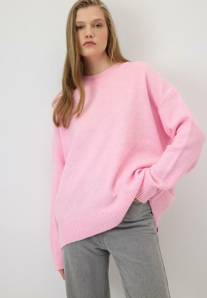 Pull oversize rose clair en maille douce, avec un ourlet et des poignets côtelés, associé à un pantalon gris.