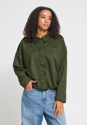 Jeune femme aux cheveux bouclés porte une veste verte oversize et un jean bleu clair, posant avec une main dans la poche sur fond blanc.