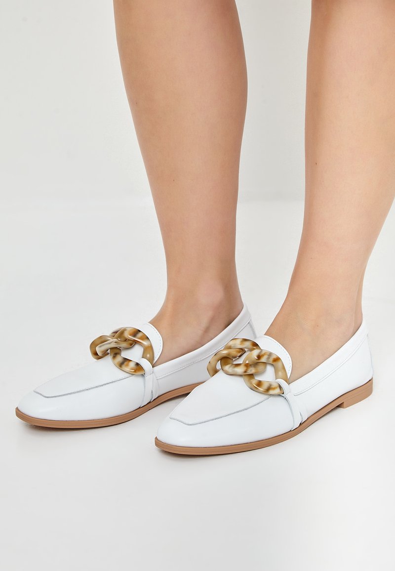 White loafers with a flat brown sole, featuring a decorative chain accent in a tortoiseshell pattern. Smooth leather upper with pointed toe design.