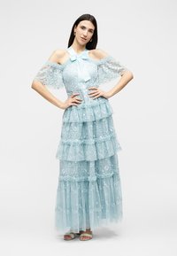 Γυναίκα που φοράει ένα ανοιχτό μπλε φόρεμα με δαντέλα σε επίπεδα, με μανίκια τύπου «κρύο ώμο» και φιόγκο στον λαιμό, στέκεται με τα χέρια στη μέση μπροστά σε απλό φόντο.