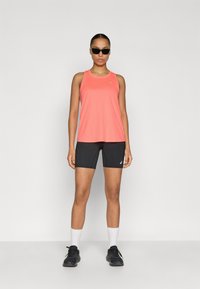 Κοραλί αθλητικό τοπ με σχέδιο racerback, σε συνδυασμό με μαύρο σορτς. Το μοντέλο φοράει μαύρα γυαλιά ηλίου και αθλητικά παπούτσια με λευκές κάλτσες.