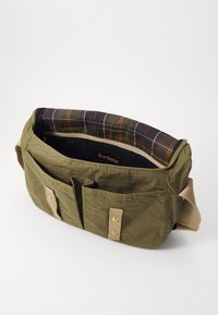 Olivově zelená messenger taška se dvěma předními kapsami, zlatým kováním a kostkovanou podšívkou. Obsahuje odolnou tkaninu a nastavitelný popruh.