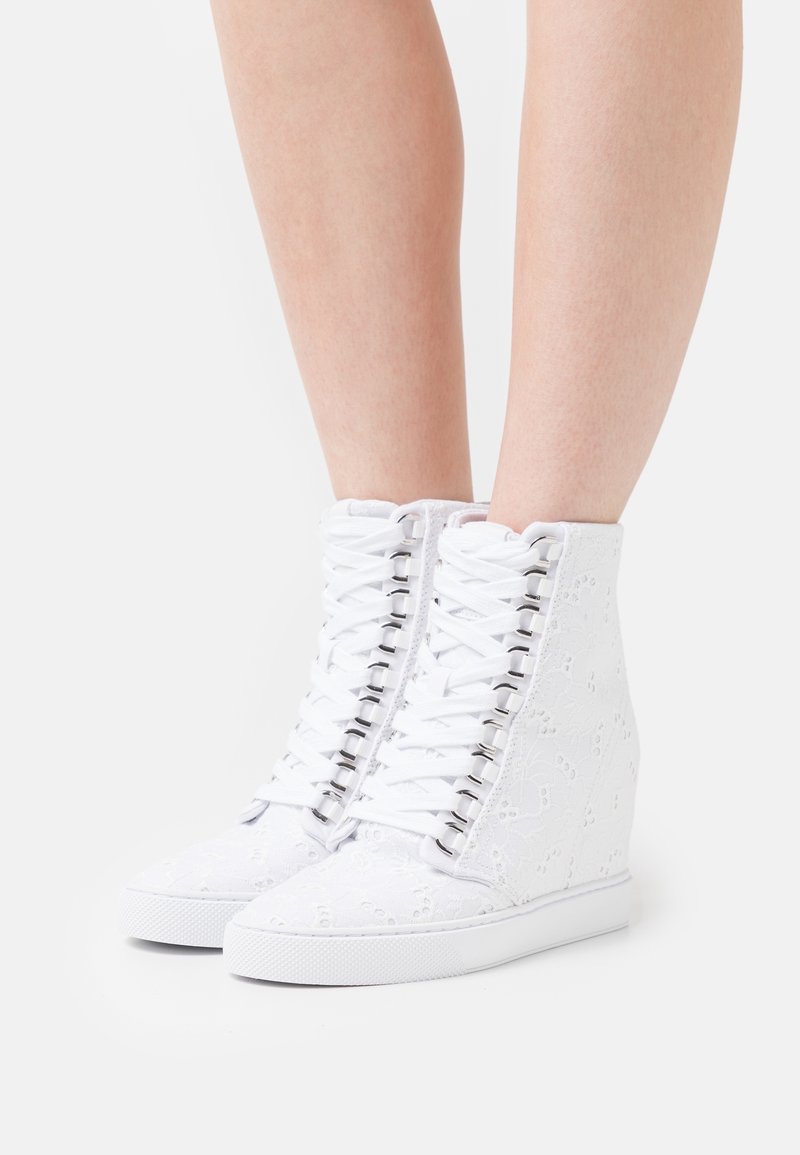 Baskets compensées blanches à lacets avec broderie florale, portées sur des jambes nues, sur un fond blanc uni.