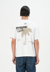 White short-sleeve t-shirt featuring a palm tree graphic and "Riverside County" text in black and beige. Casual, relaxed fit.