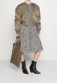 Oliivinvihreä pommittaja-takki, mustat nilkkasaappaat, kuvioitu mustavalkoinen mekko ja siihen sopiva oliivinvihreä käsilaukku. Sileät tekstuurit ja rakenteelliset muotoilut.