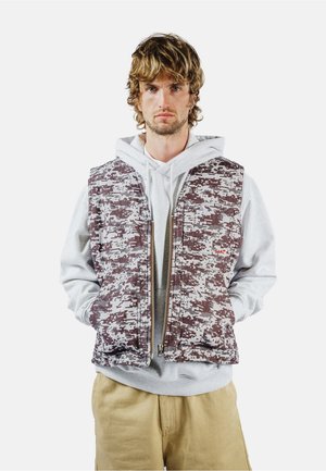 Giovane uomo con capelli mossi che indossa una felpa grigia con cappuccio, un giubbotto mimetico digitale e pantaloni beige, con le mani in tasca, sfondo neutro.