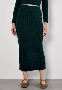 Strickmidirock in Dunkelgrün mit strukturiertem Muster, tailliert und mit gerippter Detailgestaltung, kombiniert mit braunen Mary Jane Schuhen.