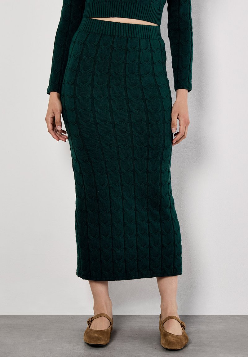 Strickmidirock in Dunkelgrün mit strukturiertem Muster, tailliert und mit gerippter Detailgestaltung, kombiniert mit braunen Mary Jane Schuhen.