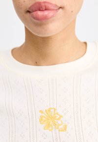 Crème kleurig top met textuurpatronen, voorzien van verticale strepen en een gele bloemenborduursel.