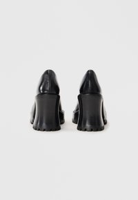 Ζευγάρι μαύρα δερμάτινα παπούτσ�ια με χοντρό τακούνι και σόλες με πέλματα, απεικονιζόμενα από πίσω σε απλό λευκό φόντο.