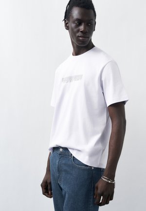Man wearing white Calvin Klein t-shirt and blue jeans, looking to the side against a plain light background.