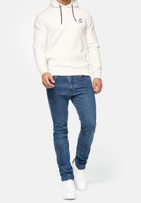 Hoodie crème avec des cordons noirs et un logo, associé à un jean slim bleu et des baskets blanches. Texture lisse, design décontracté.