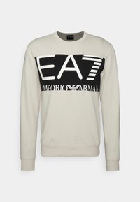 Ljusbeige sweatshirt med en svart horisontell panel som har vit text "EA7 EMPORIO ARMANI." Tillverkad av bomull med långa ärmar och ribbat muddar.