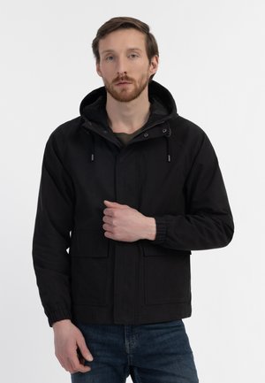 Veste noire avec capuche, poches avant, poignets élastiques et fermeture éclair. Le modèle se tient contre un fond blanc uni.