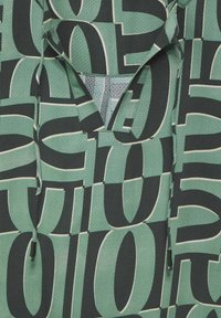 Sudadera verde con un patrón, de cuello en V, que presenta una mezcla de letras en negrita de diferentes tamaños y texturas, hecha de un material ligero.