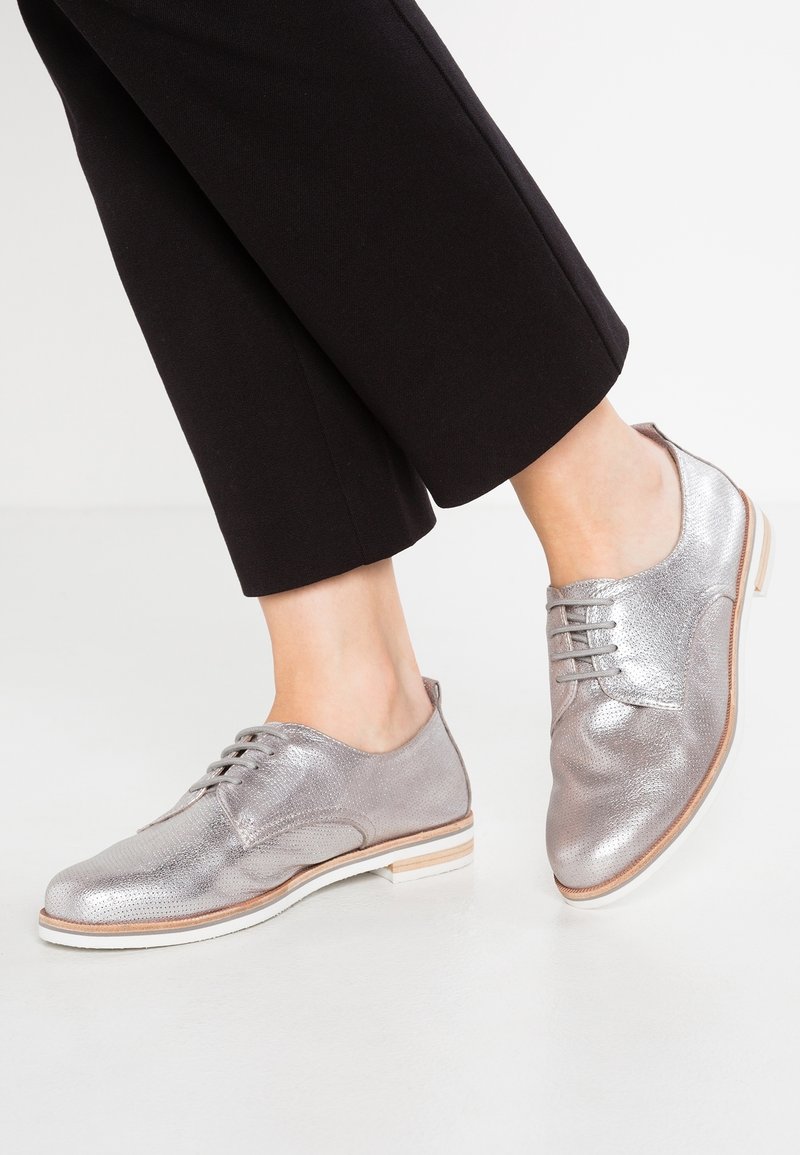 Chaussures montantes en cuir argenté texturé, avec bout rond, semelle blanche et accents bruns subtils, portées avec un pantalon noir raccourci.