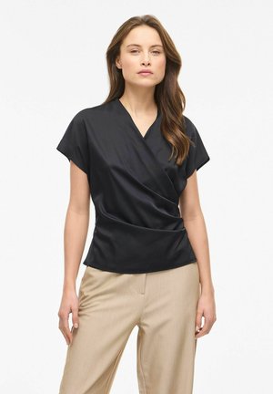 Femme portant un chemisier cache-cœur noir à manches courtes et un pantalon beige, debout devant un fond blanc uni.