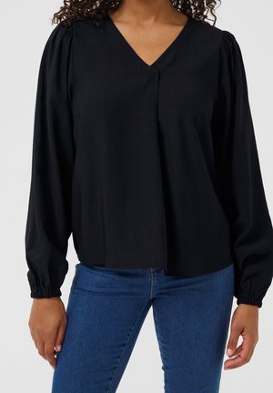 Frau trägt eine schwarze lange V-Ausschnitt-Bluse mit gerafften Bündchen und blaue Jeans vor einem einfarbigen Hintergrund.