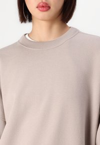 Žena v béžovém svetr s kulatým výstřihem a velkými náušnicemi na kruhu, s rovnými vlasy do délky ramen a lesklými růžovými rty.