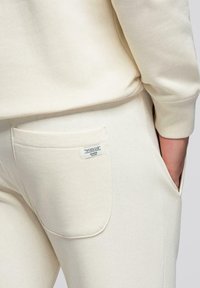 Gros plan sur une personne portant un pantalon de survêtement crème avec une poche arrière et une manche de sweat assortie visible.