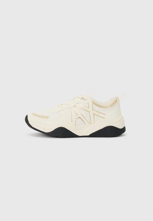 Niskie beżowo-białe sneakersy z czarną podeszwą i dużym logo "AX" po boku, widziane z zewnętrznej strony na neutralnym tle.
