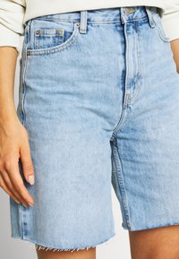 Pantalones cortos de mezclilla azul claro con dobladillo deshilachado, con bolsillos frontales, cierre de botón y un ajuste ligeramente holgado.