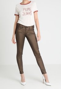 Camiseta blanca de manga corta con ribetes rojos, que presenta el texto "Pepe Jeans". Jeans ajustados de bronce brillante. Tacones blancos completan el conjunto.
