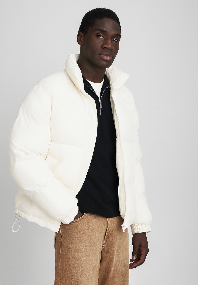Giacca imbottita crema, con colletto alto, chiusura con zip e polsini elasticizzati. Indossata sopra un top nero e pantaloni beige. Tessuto morbido.