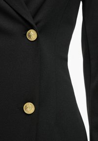 Blazer noir cintré avec deux boutons décoratifs dorés présentant des motifs embossés complexes.