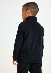 Pullover in pile nero con colletto alto, superficie texturizzata e maniche lunghe, indossato da un bambino che guarda leggermente lontano dalla fotocamera.