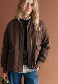 Blonde vrouw met een bruine doorgestikte jas over een gestreept shirt en trui, die een tas met groenten vasthoudt, tegen een bruine achtergrond.