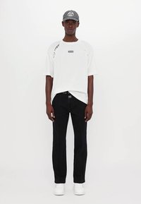 T-shirt blanc à manches courtes avec des détails graphiques, associé à un jean noir avec des coutures contrastées, et des baskets blanches. Une casquette grise complète le look.