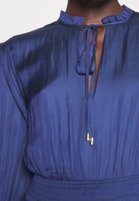 Vestido de satén azul marino con un cuello alto con volantes, apertura en forma de ojal y cordón ajustable con herrajes dorados. Detalle de cintura fruncida. Mangas largas.