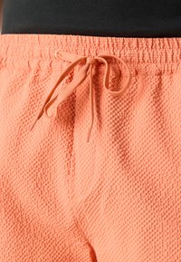 Gros plan sur un short en tissu velouté couleur pêche avec une taille élastique et un cordon de serrage noué, porté avec un haut noir visible au-dessus de la taille.