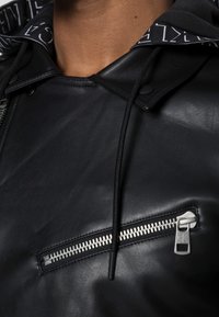 Giacca di pelle nera con tasca sul petto con zip, cappuccio regolabile con bordino logo e texture liscia con design aderente.