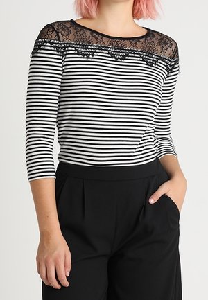 Vrouw draagt een zwart-wit gestreepte top met een zwarte kanten halslijn en zwarte hoge taillebroek, poseert met één hand in haar zak.