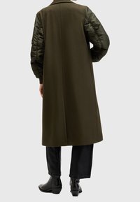 Abrigo verde oliva de diseño largo y recto, combinado con mangas acolchadas oscuras. Cuenta con cuello clásico y abertura lateral para facilitar el movimiento.