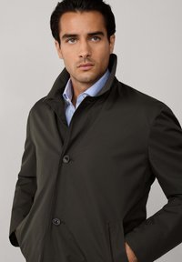 Tummanvihreä, napitettava takki, jossa on korkea kaulus, sileä pinta ja sivutaskut. Alla on näkyvissä vaaleansinisesti raidoitettu paita.